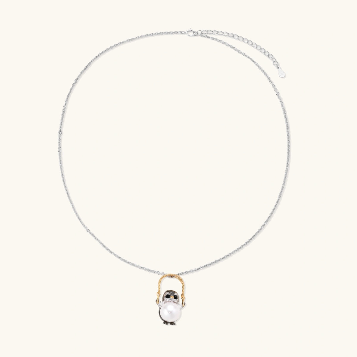 Whimsical owl pendant holding a white pearl on a gold swing, rhodium-plated silver chain on cream background.