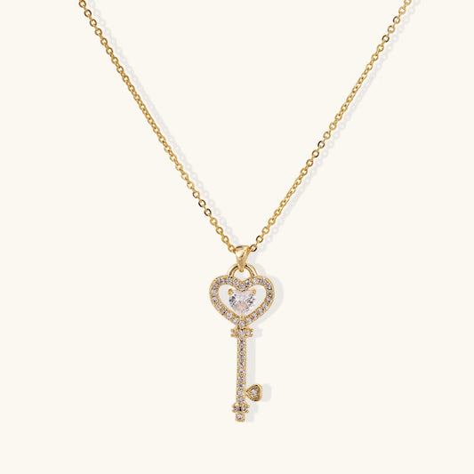 Delicate gold key pendant with heart-shaped bow and crystal pavé detailing on fine chain against cream background.