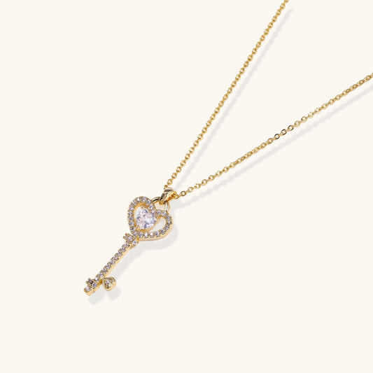 Delicate gold key pendant with heart-shaped bow and crystal pavΓ© detailing on fine chain against cream background.