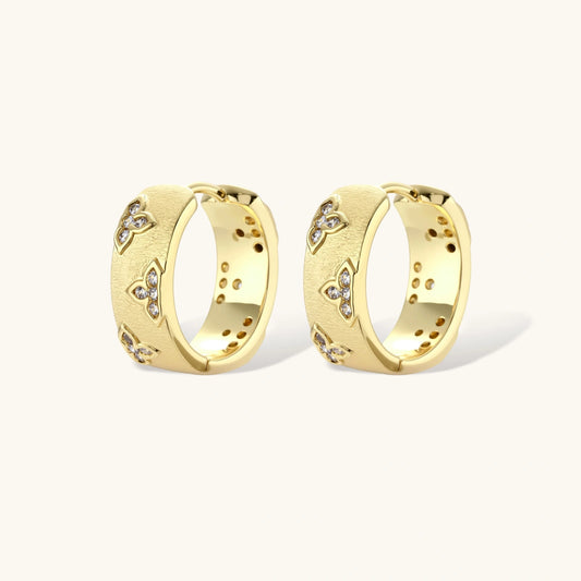 Gold-plated hoop earrings with textured surface and raised floral motifs set with clear zirconia stones on cream background.