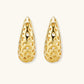 Close-up of small gold-plated hoop earrings with intricate honeycomb cutout pattern on cream background.