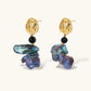 Ornate gold-plated drop earrings with black beads and iridescent blue-purple baroque pearls against white background.